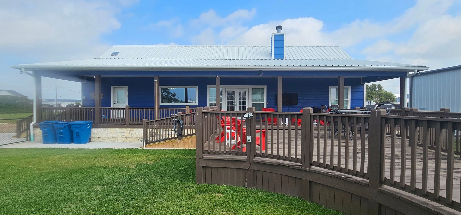 A blue, single-story house with a white metal roof, a large wooden deck, and blue trash bins on a sunny day.
