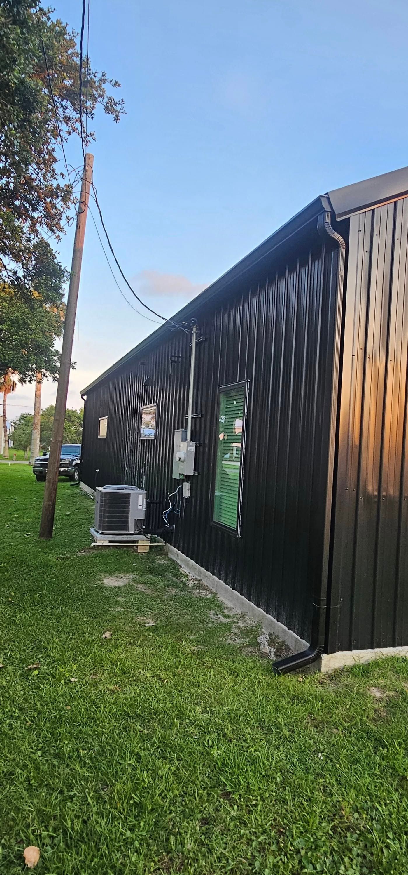 Side view of a black metal building with an HVAC unit, electrical service, and utility pole on a grassy lawn.