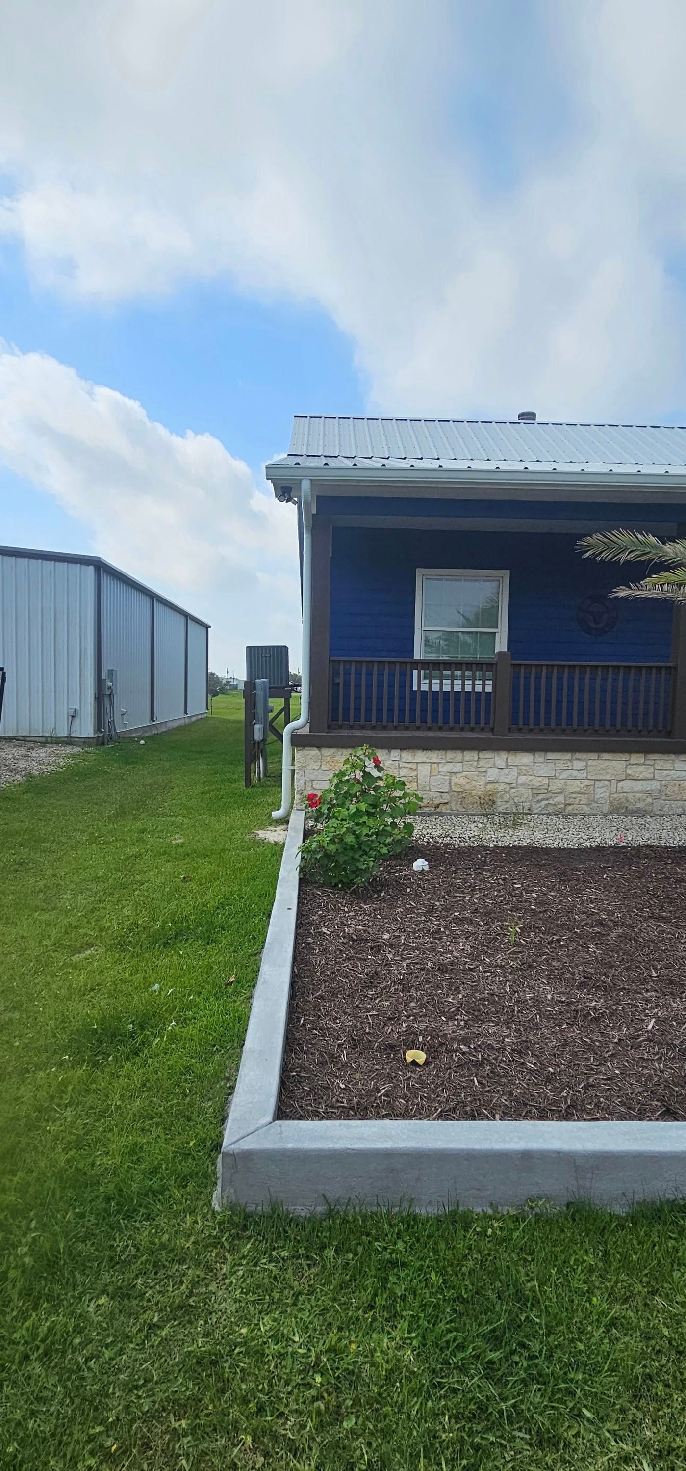 A blue house with a front porch and stone base sits next to a metal outbuilding under a bright, cloudy sky.