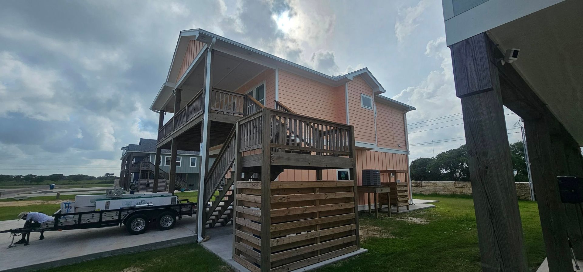 An elevated, peach-colored beach house with a wooden staircase and privacy lattice stands on a sunny, grassy lot.