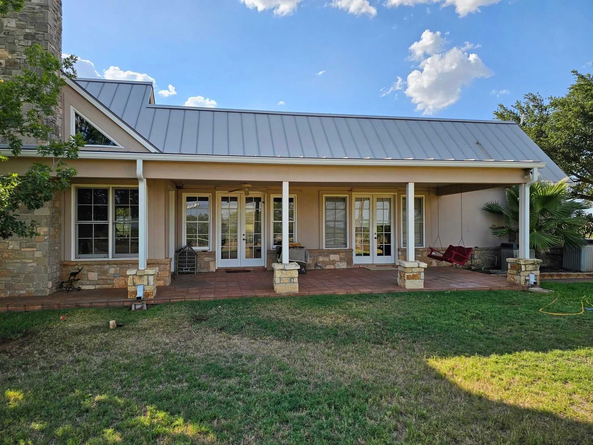 A tan house with a metal roof and a spacious, covered stone patio featuring two sets of double glass doors.