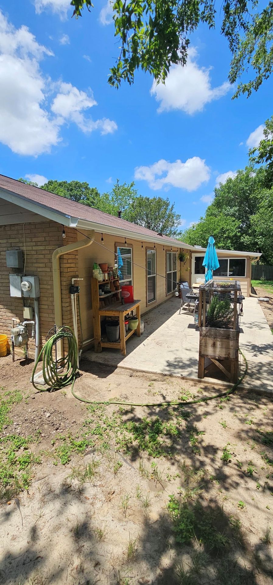A tan brick house with a covered patio, a wooden workbench, a potted plant, and a blue umbrella on a sunny, clear day.