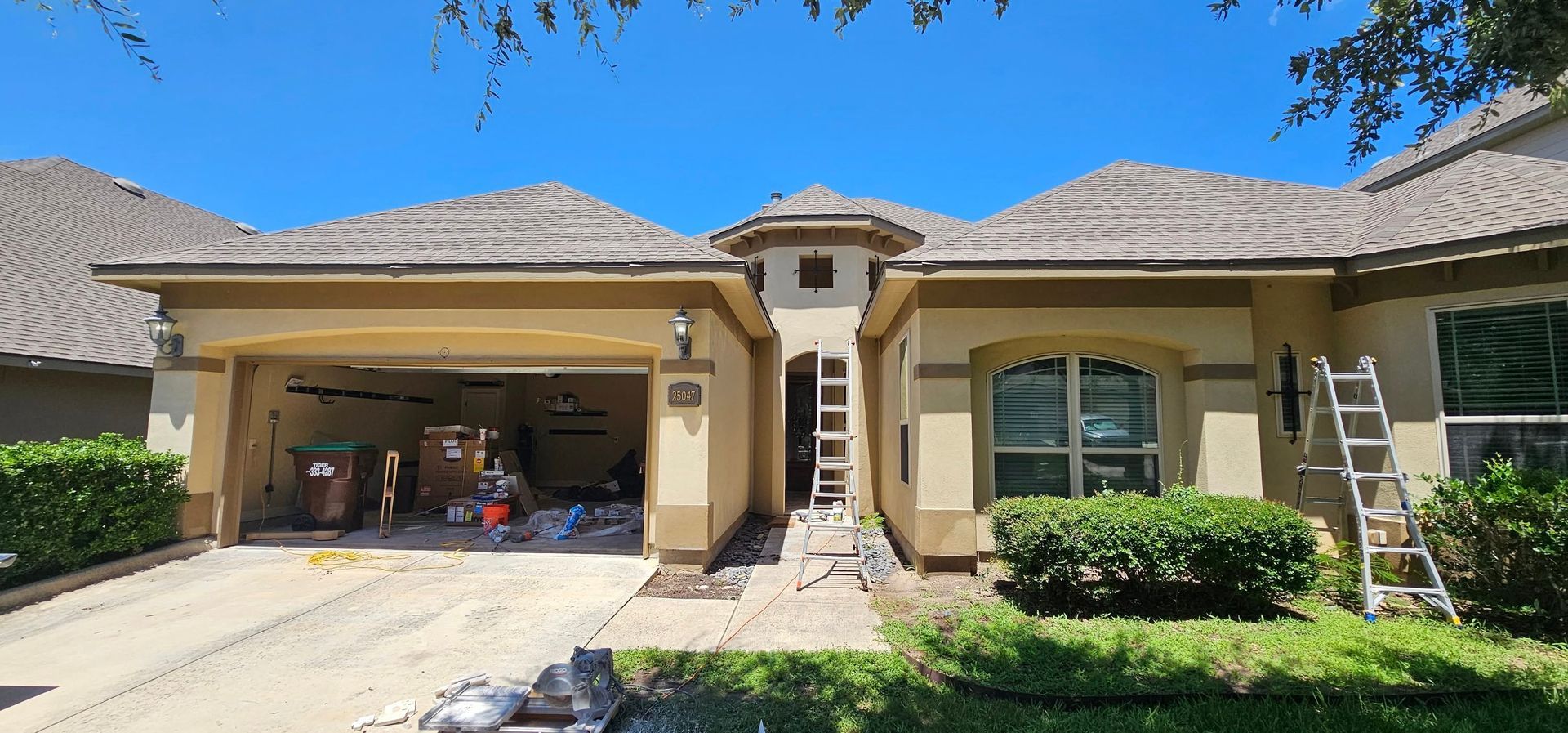 A tan, single-story suburban house with a garage, tiled roof, and a ladder placed near the center entrance.
