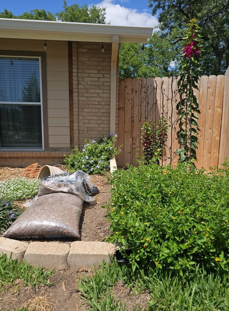 A house corner with a stone-brick wall, a wooden fence, a blooming purple hollyhock, and a bag of mulch in the garden.