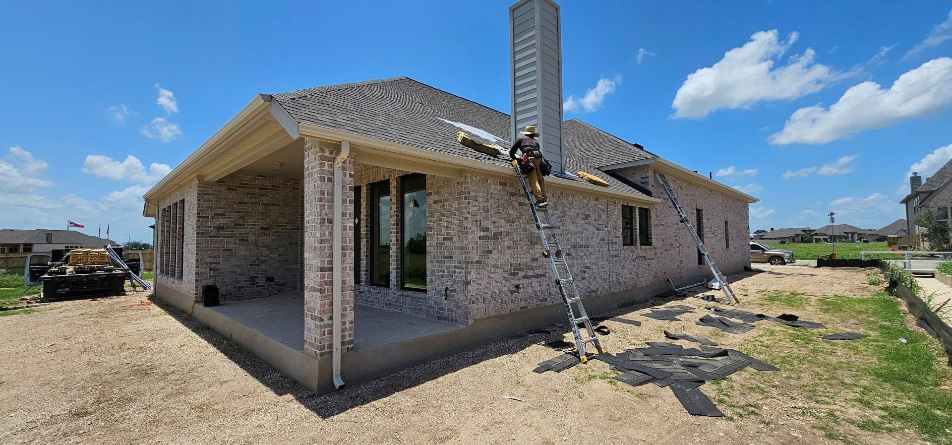 A person on a ladder repairs the roof of a beige brick house under a blue sky.
