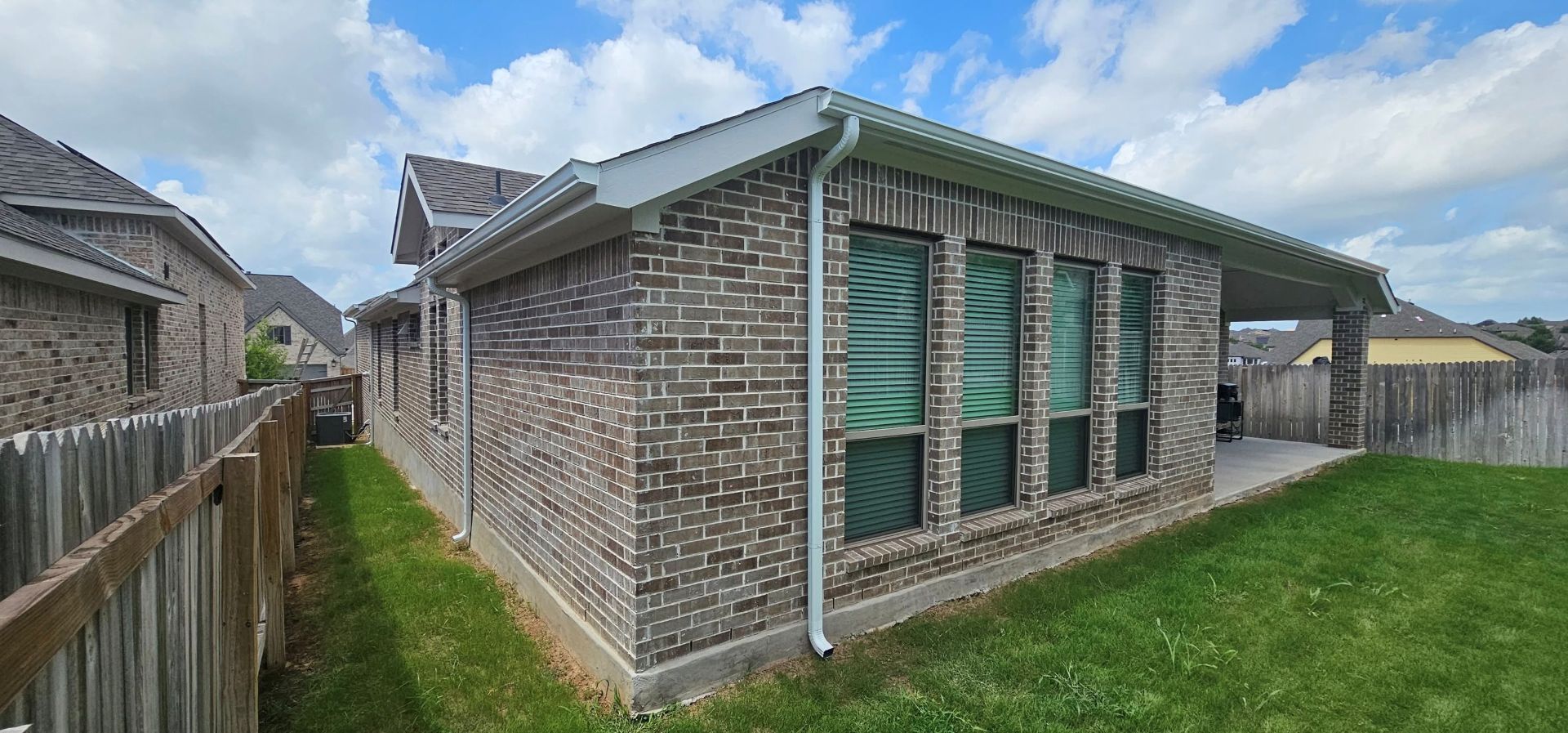 The back exterior of a single-story brick house with a covered patio, grass lawn, and a wooden fence.