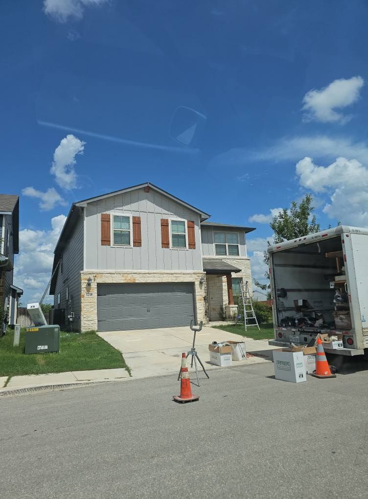 A two-story gray house with a stone base and orange shutters stands by a road with a moving truck parked in the driveway.