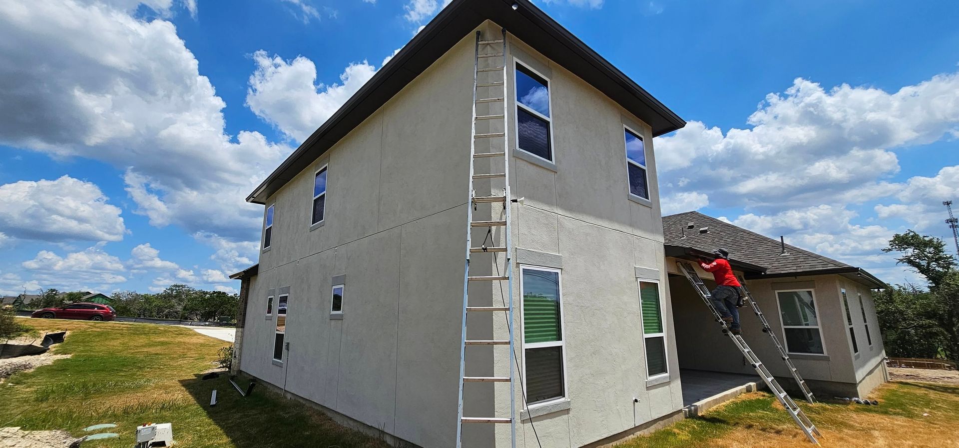 Construction workers on ladders perform stucco exterior work on a two-story residential house under a sunny blue sky.