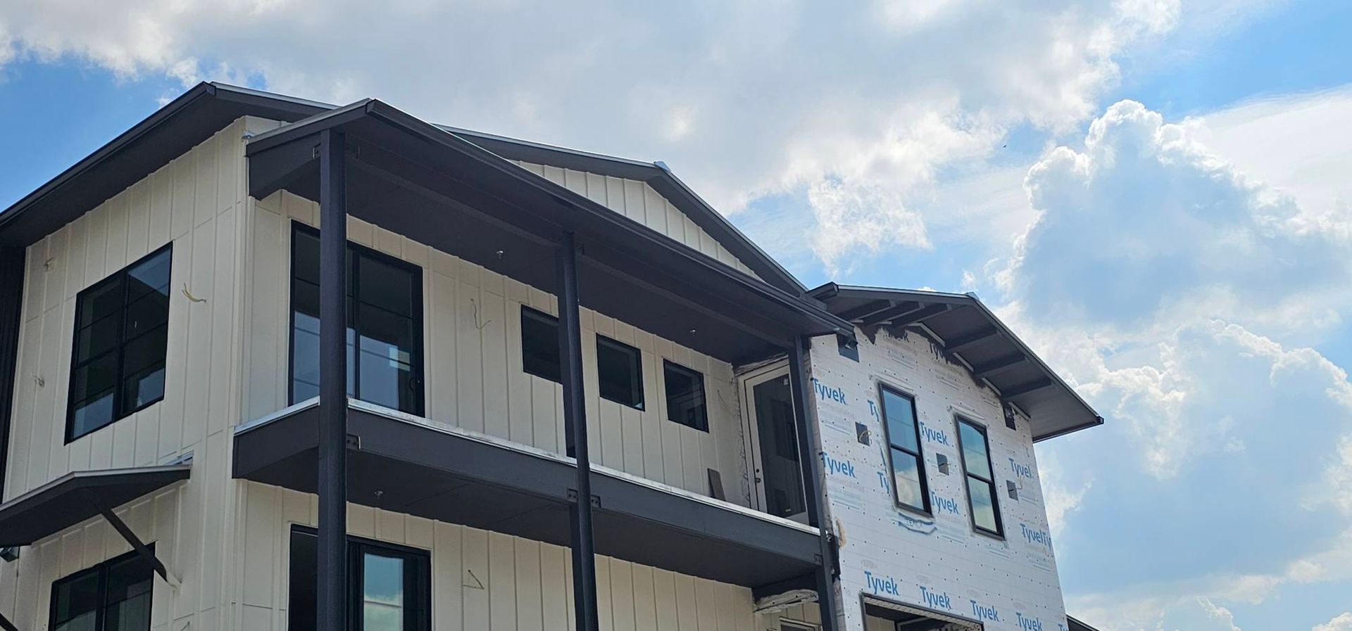 A multi-story house under construction with white exterior sheathing, black window frames, and a dark roof under blue sky.