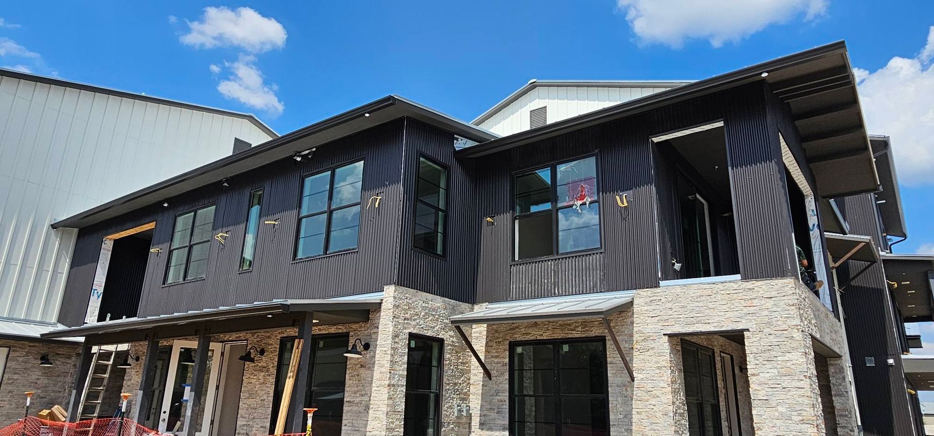 A two-story building under construction with dark metal siding, light-colored stone veneer, and large windows.