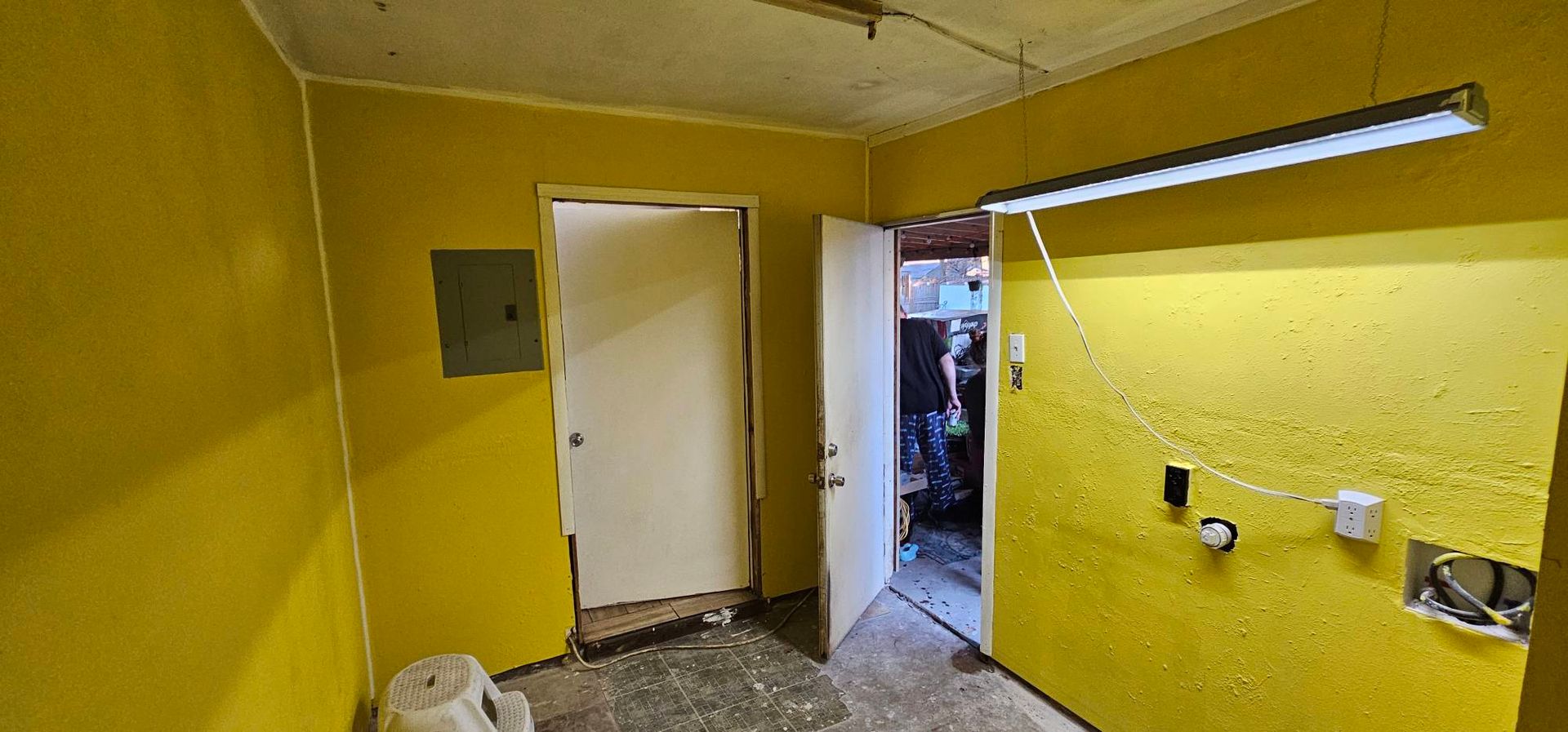 A small room with bright yellow walls, concrete flooring, two doorways, a breaker box, and an overhead fluorescent light.