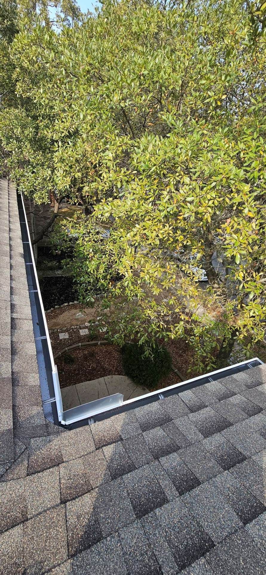 A high-angle view of a roof valley with light-colored metal flashing, bordered by textured gray shingles and green trees.