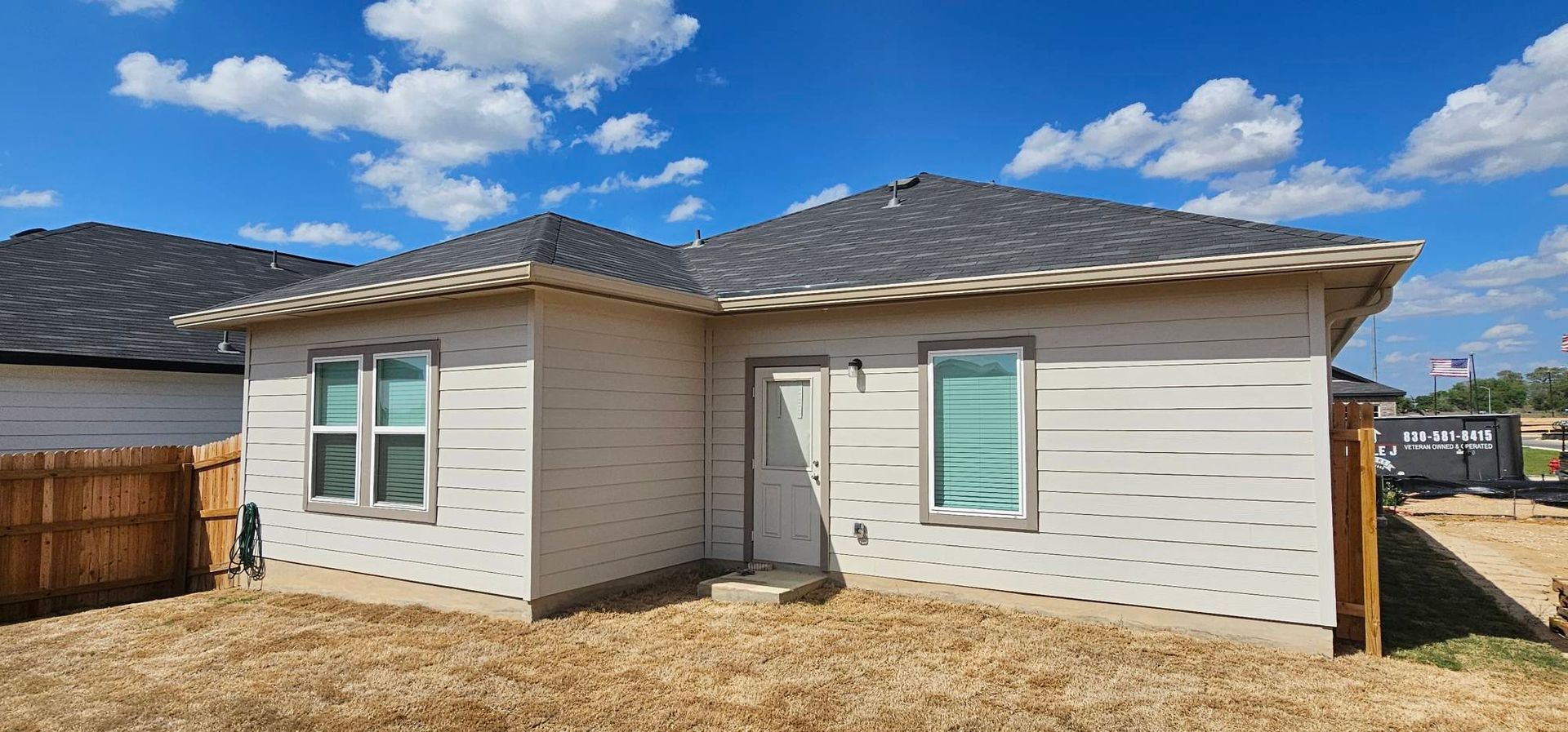 A single-story, light-colored house with a dark shingle roof, glass windows, and a side door in a fenced backyard.