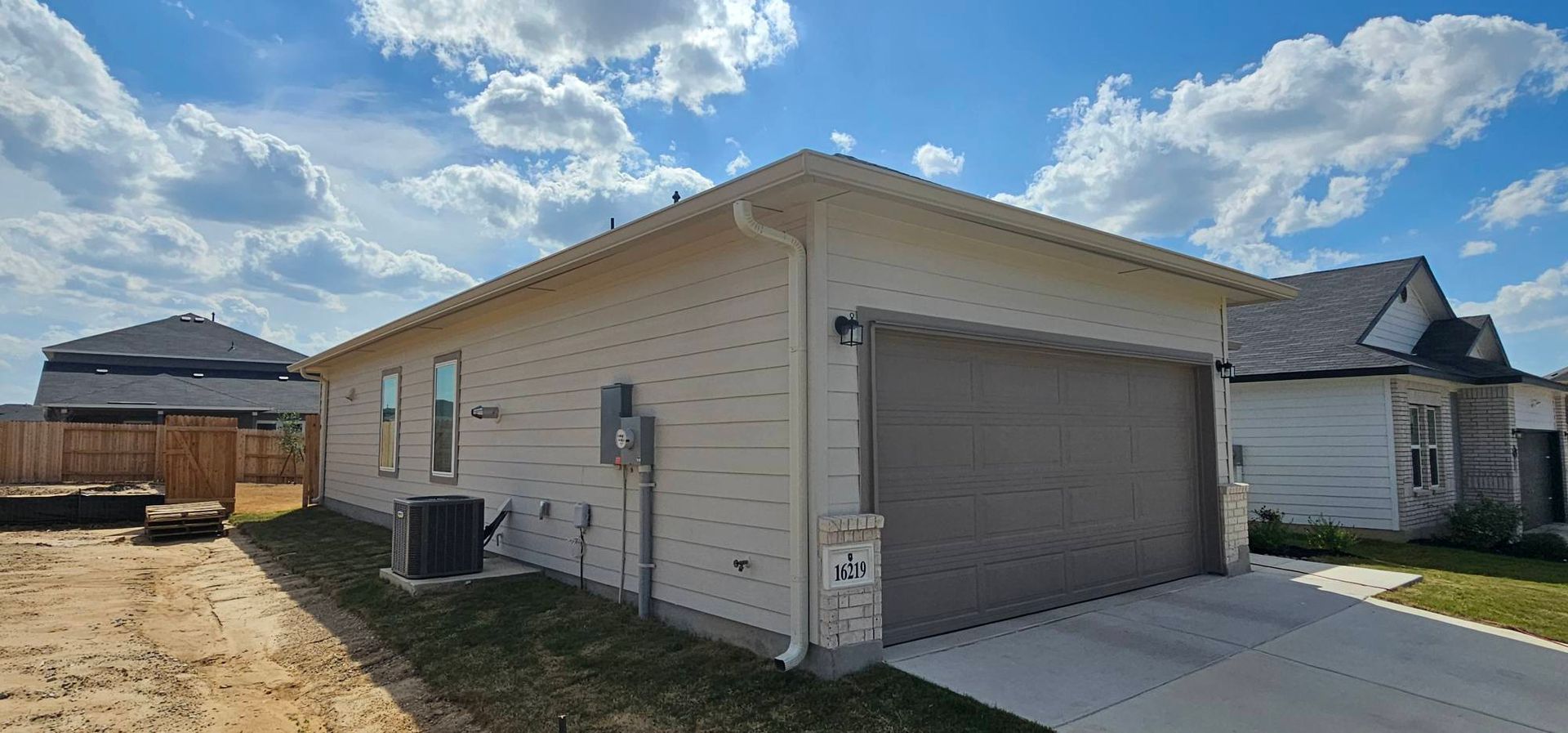 The side and garage exterior of a newly built, light-colored house with a concrete driveway on a sunny day.