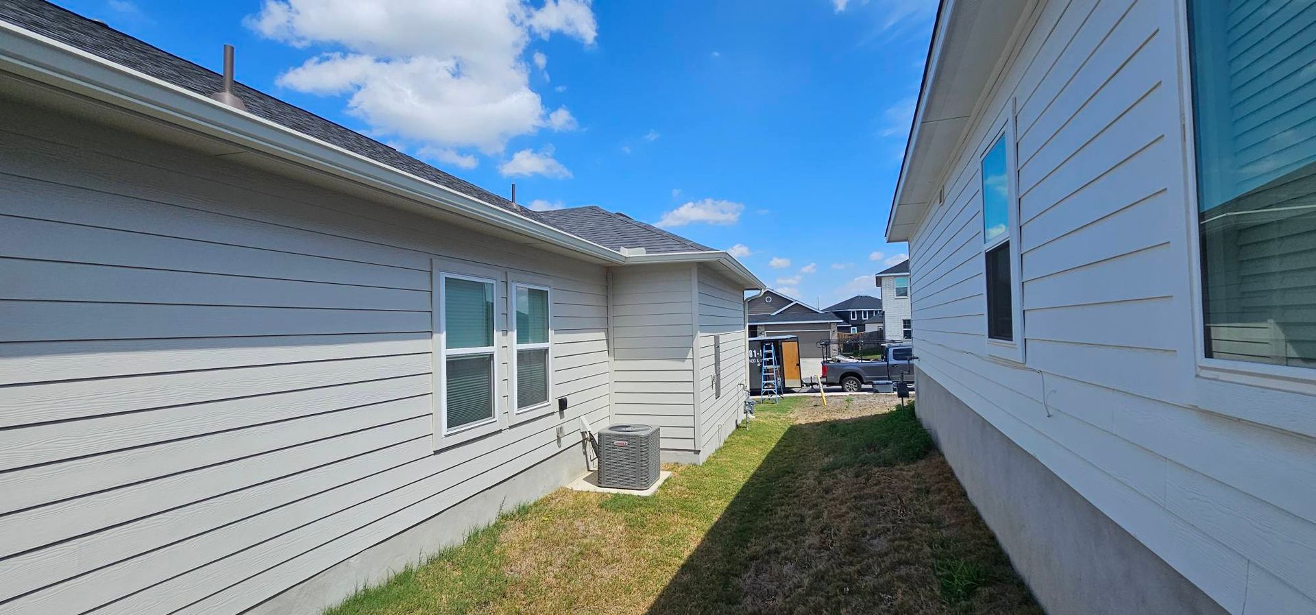 A view looking down the narrow, grassy space between two light-colored suburban houses under a bright blue sky.