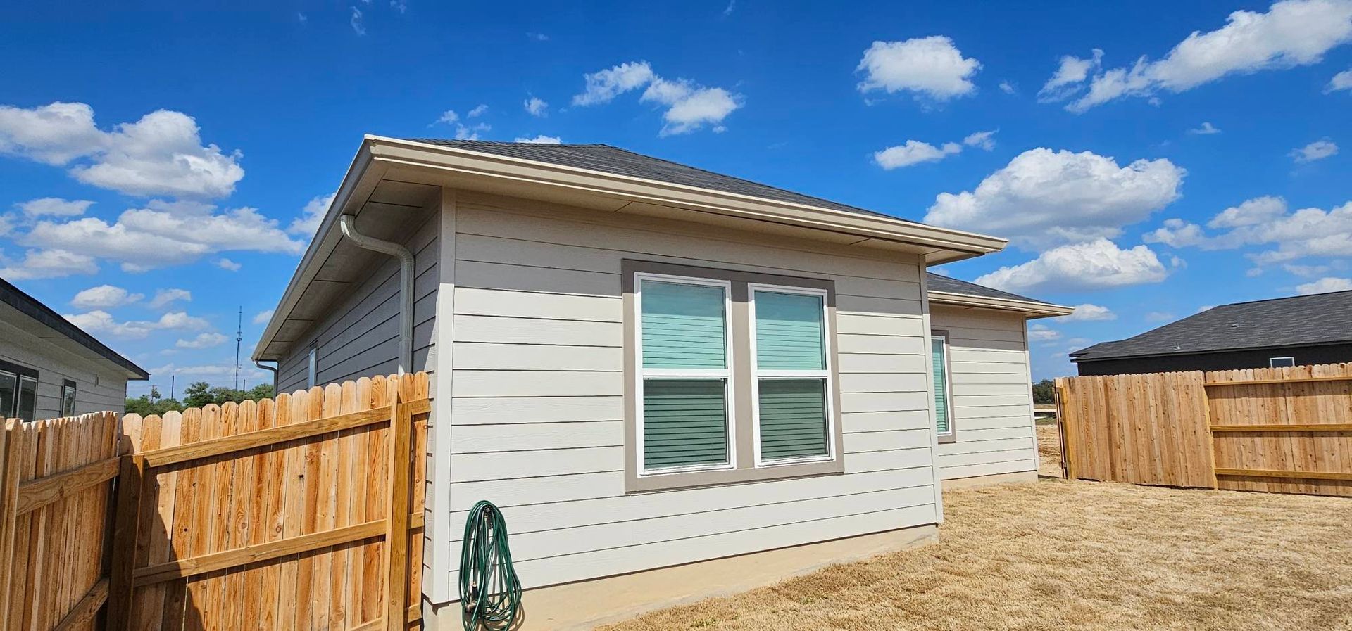 A single-story, light-colored house exterior under a blue sky with scattered clouds, viewed from a dry, grassy backyard.