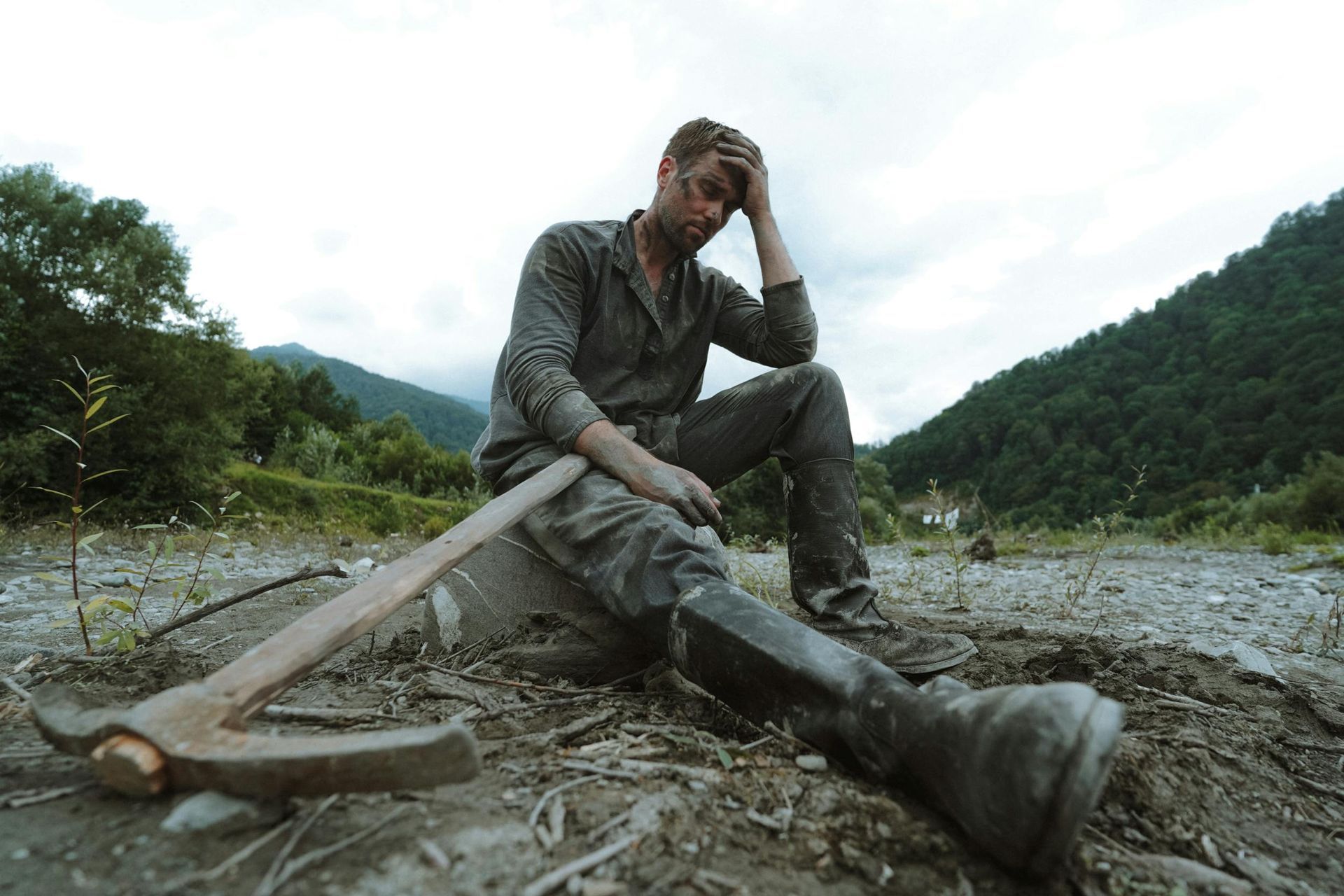 A man sitting on the ground with his head in his hand and a pick ax leaning on his lap.