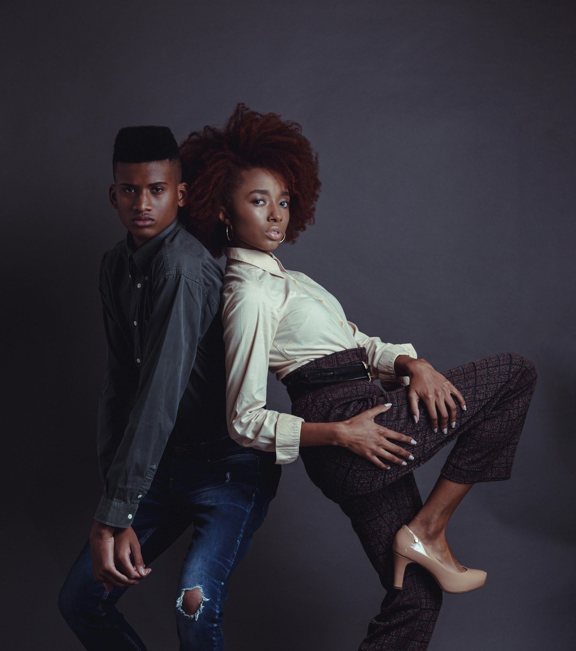 Two models, leaning back-to-back. Man in denim, woman in patterned pants. Studio, grey background.