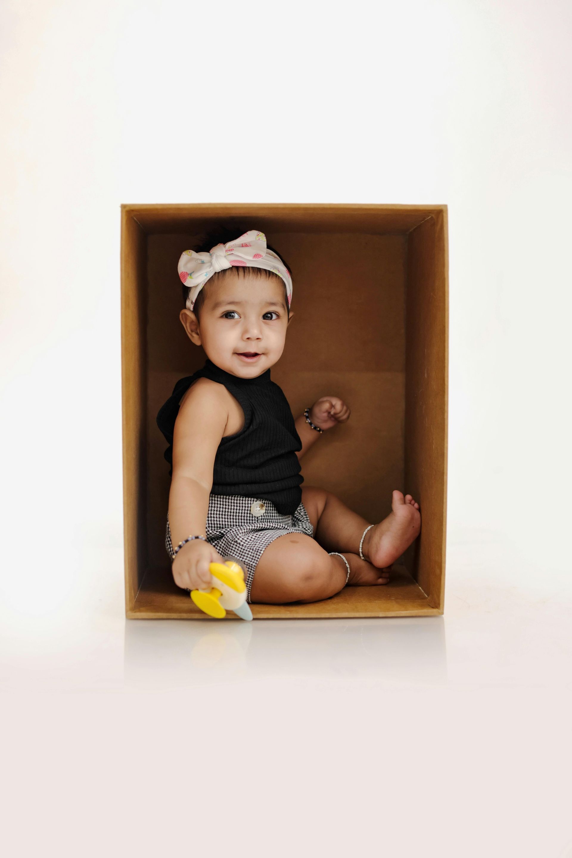 Baby in a box wearing a headband and black top smiles, holding a toy.