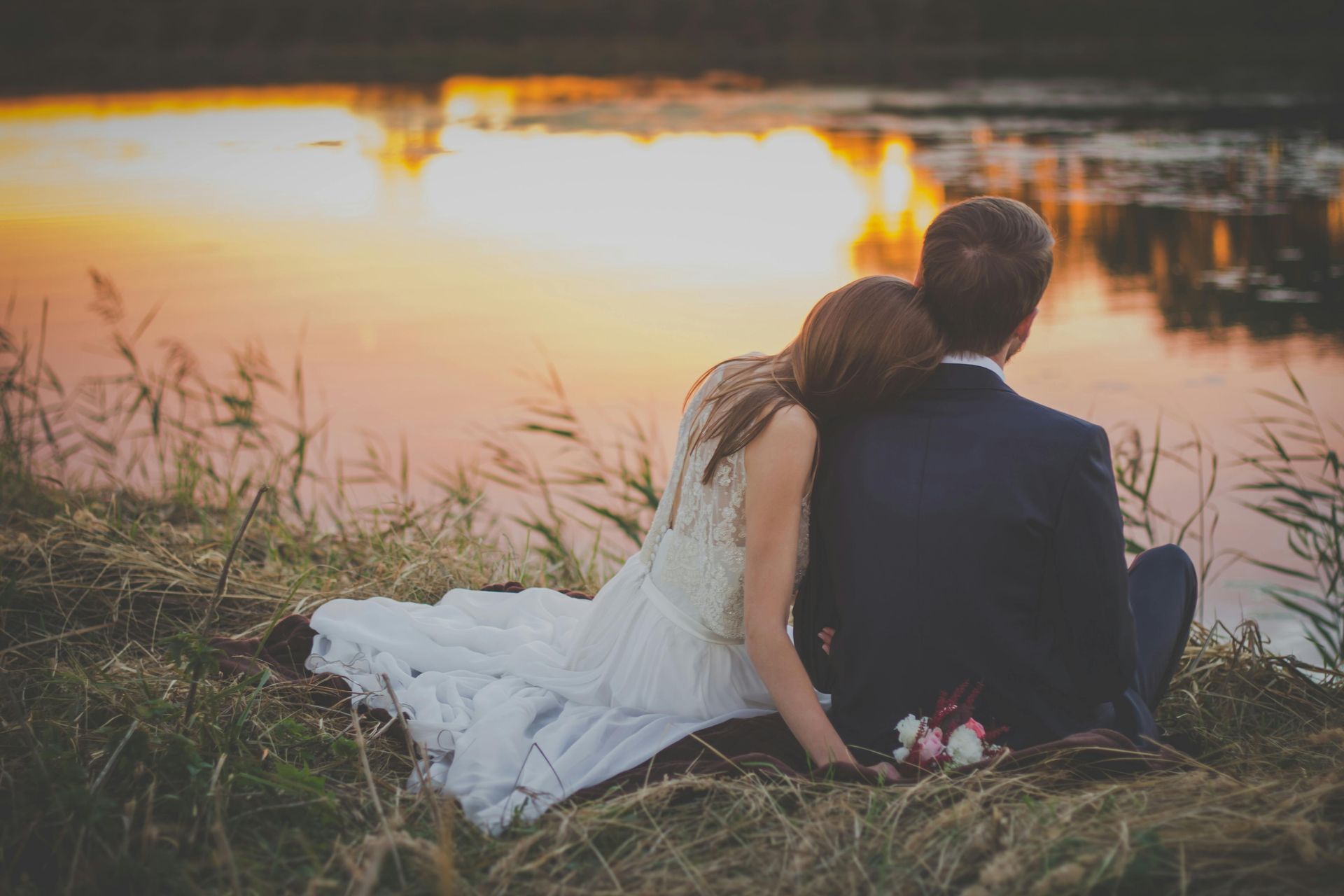 A bride and groom are sitting on the grass near a lake at sunset.