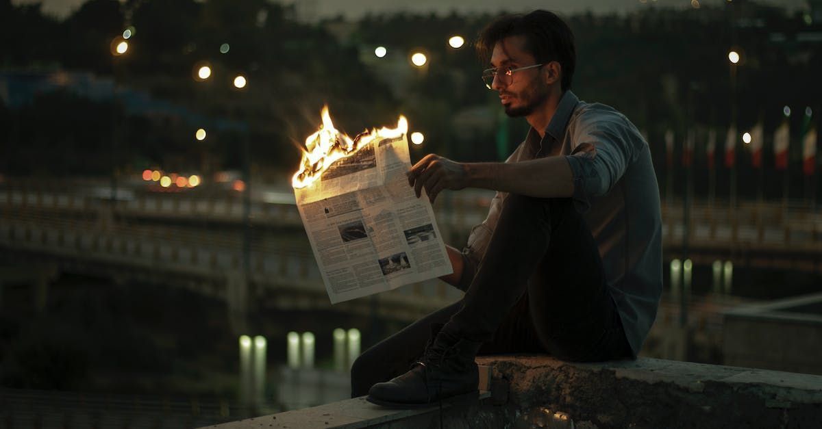 A man sitting down in front of a small bridge holding a newspaper that is on fire at the top. 