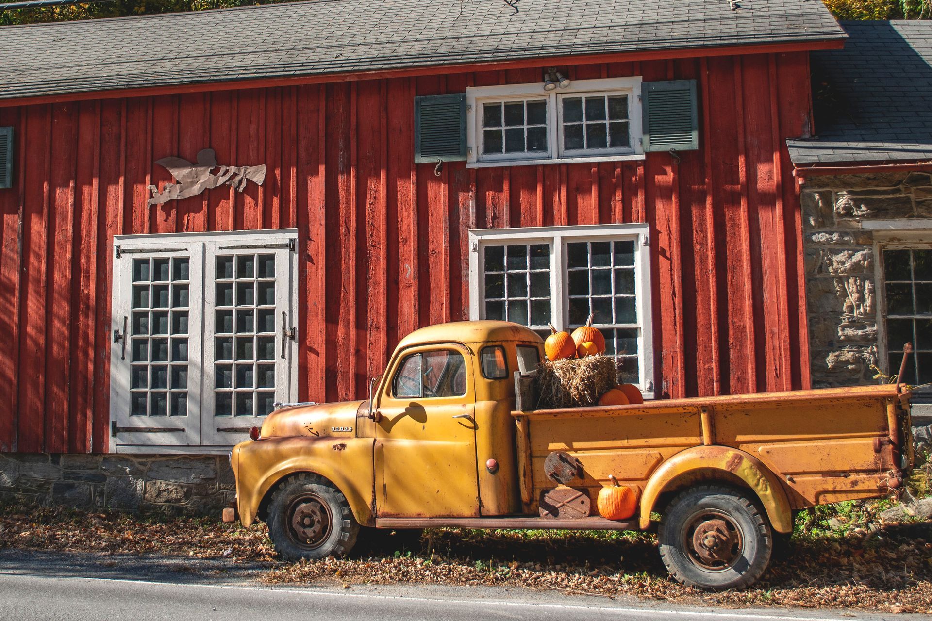 A yellow truck with pumpkins in the back is parked in front of a red barn.