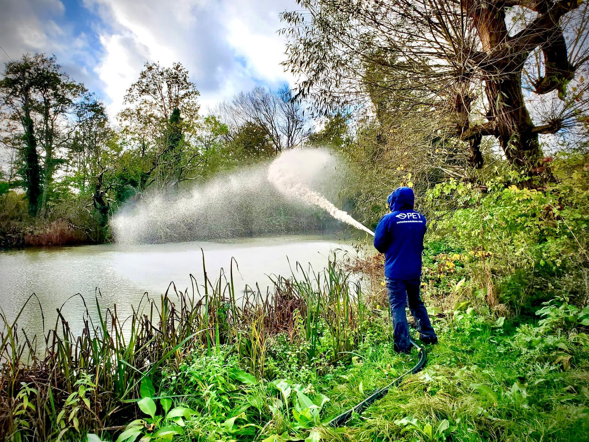 Person in blue watering plants by a lake with a spray of water, surrounded by green trees and reeds.