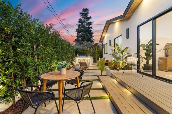 Patio de jardin accueillant au coucher du soleil avec chaises en osier, table en bois, guirlandes lumineuses et façade de maison moderne.