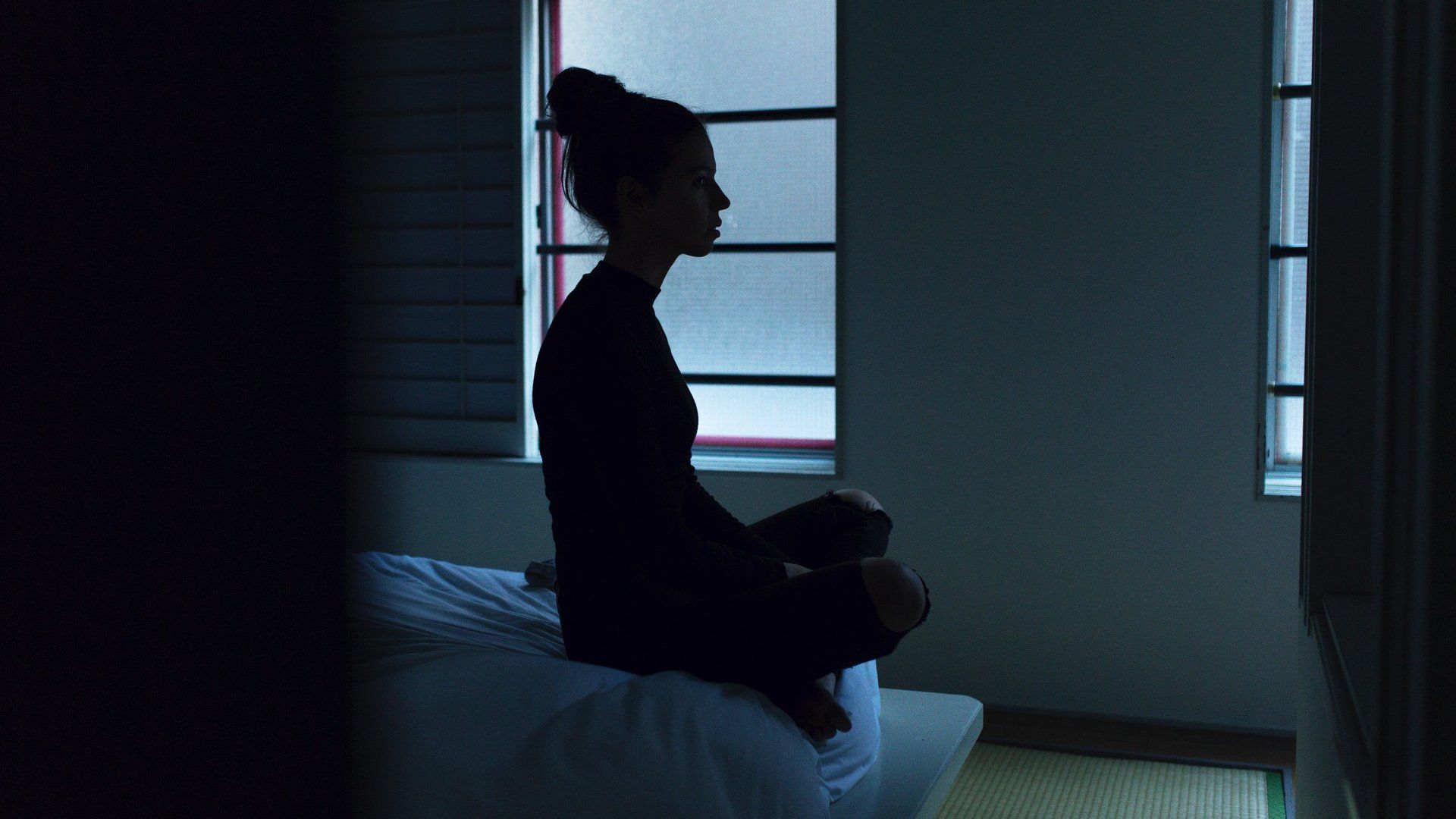 A woman is sitting on a bed in front of a window in a dark room.