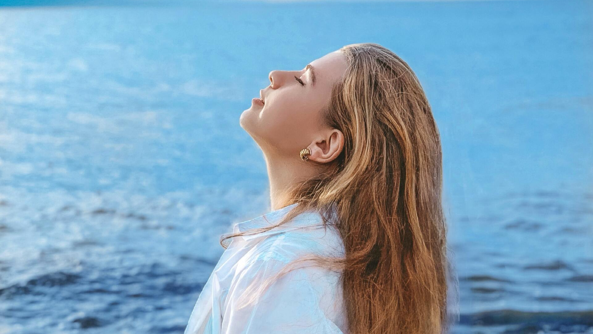 A woman is standing on a beach with her eyes closed and her hair blowing in the wind.