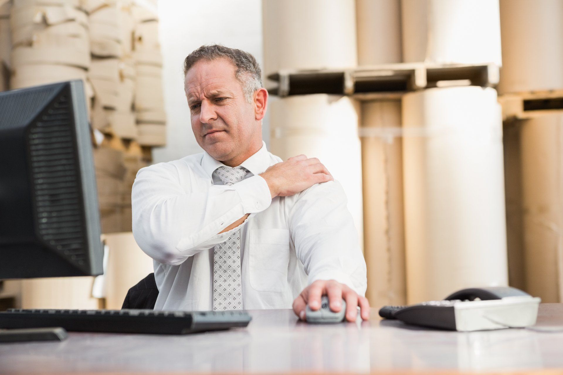 A man is sitting at a desk with a computer and holding his shoulder.