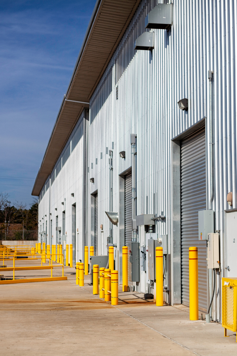 Industrial building exterior with corrugated metal siding, loading bays, and bright yellow bollards under a blue sky.
