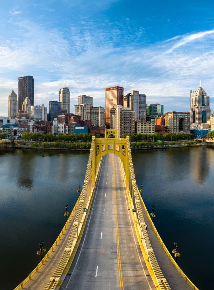 Yellow bridge over water leads to a city skyline with tall buildings under a blue sky.