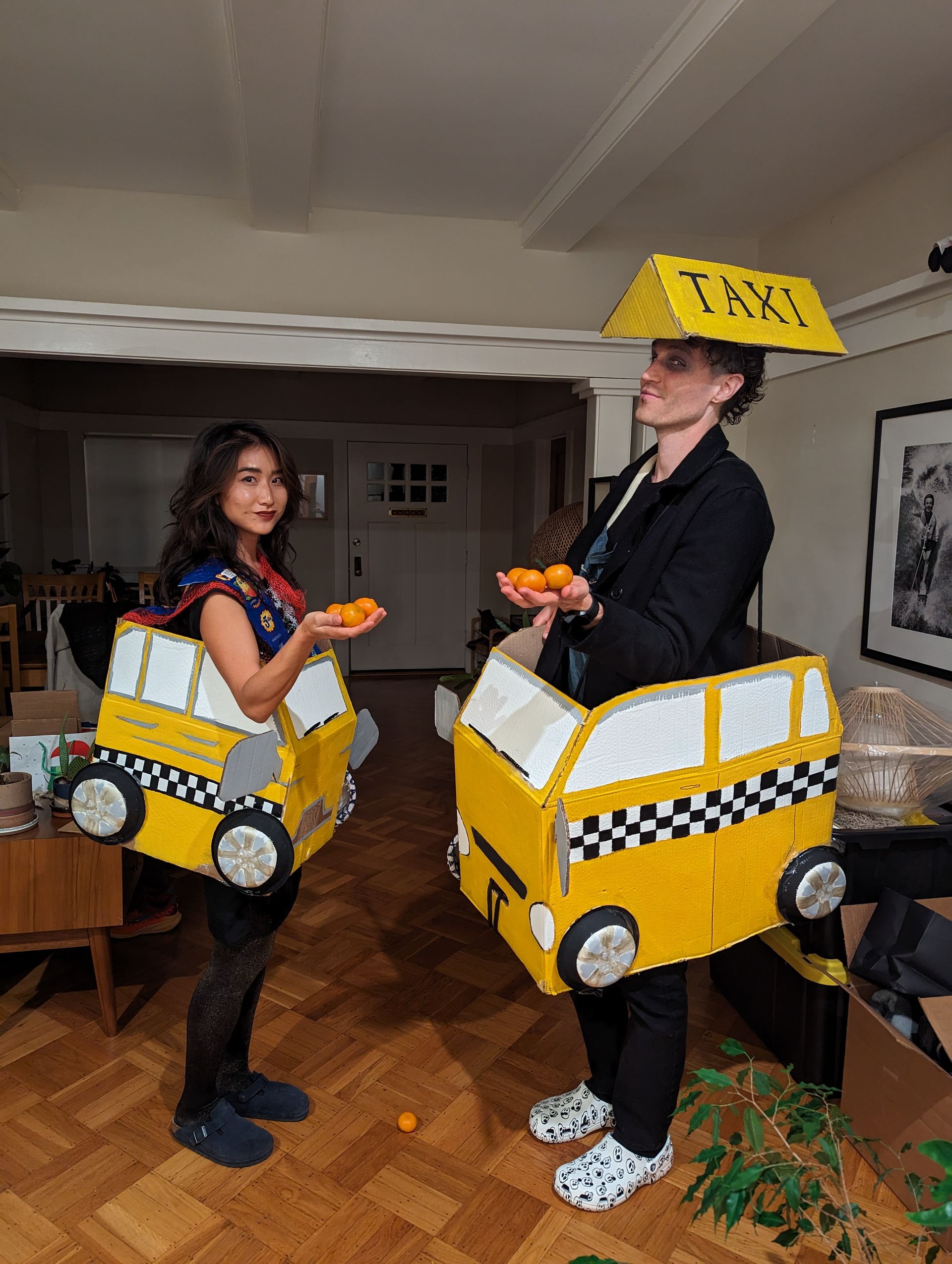Two people in homemade taxi costumes holding oranges indoors.