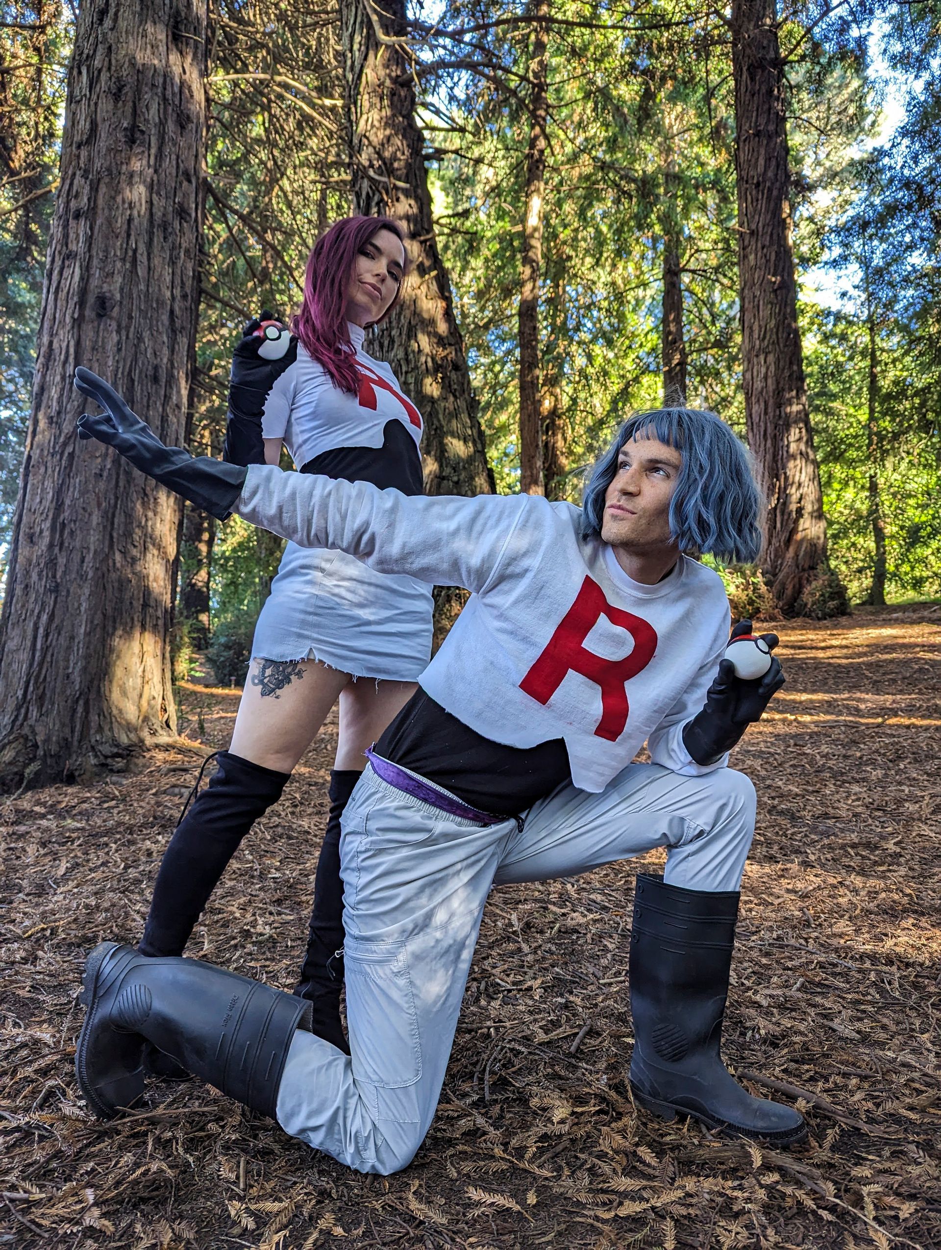 Two people dressed as Team Rocket characters pose in a wooded area. The person in front kneels, arm outstretched.