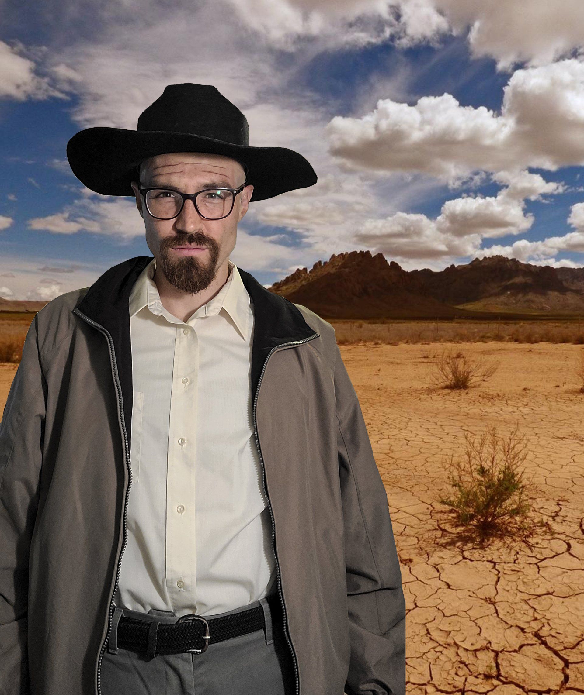 Man in hat and glasses, standing in desert, wearing a jacket and button-down shirt.