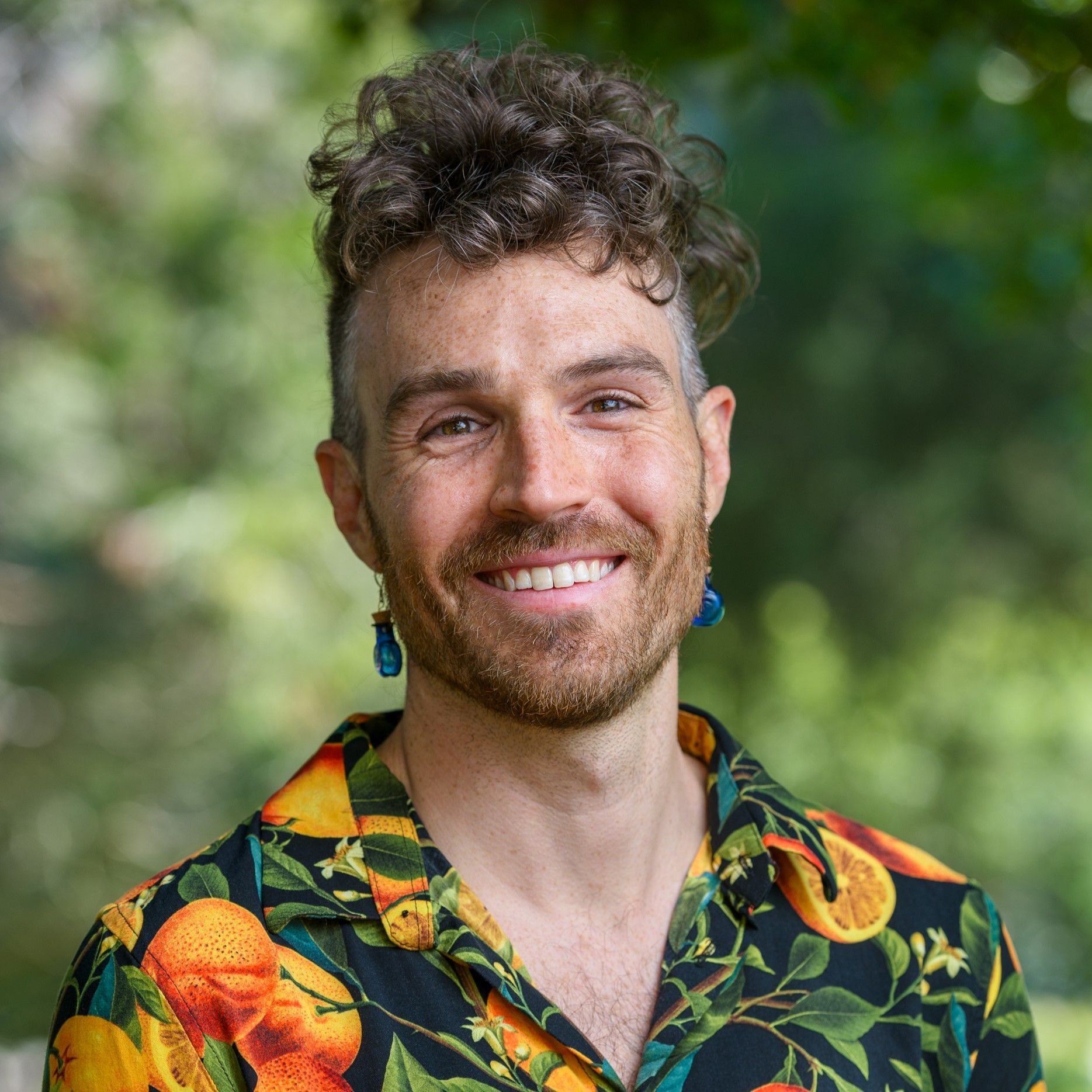 Man with curly hair and earrings smiles outdoors, wearing a tropical shirt with oranges.