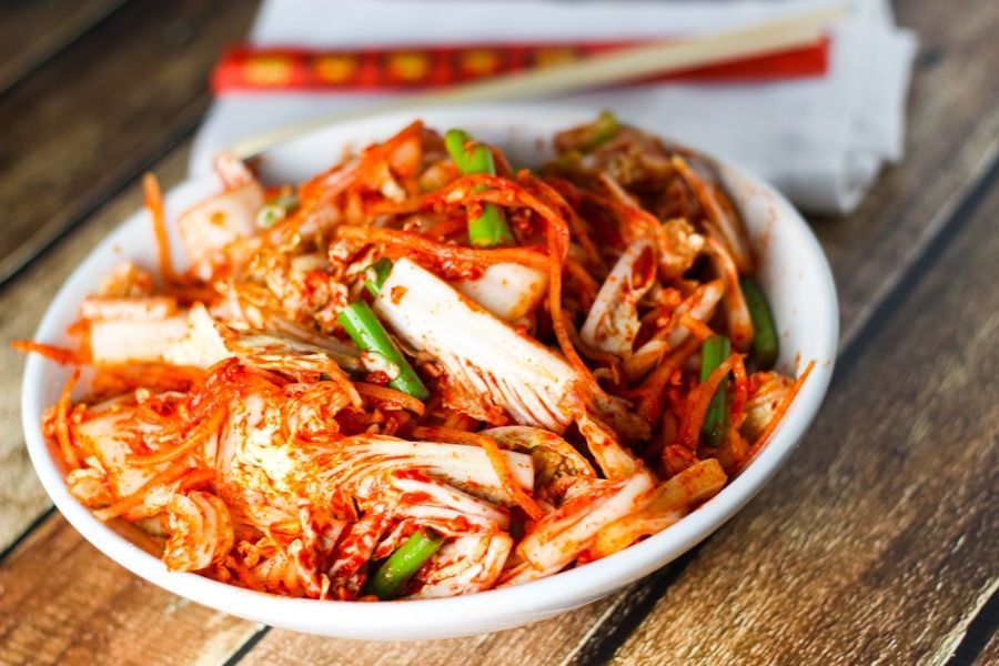 Bowl of spicy kimchi with green garnish on a wooden table, with chopsticks in the background