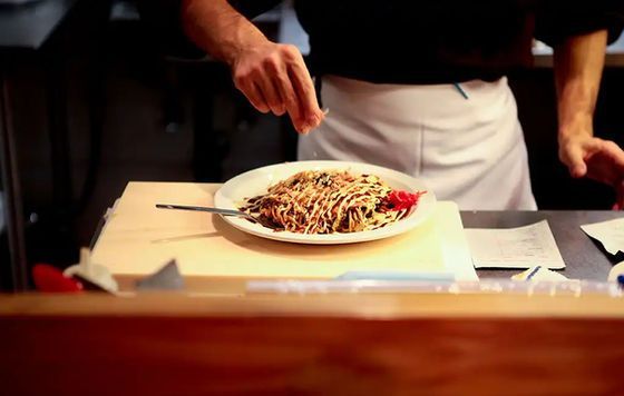 Chef plating pasta on a white plate in a restaurant kitchen, with a red garnish on the side