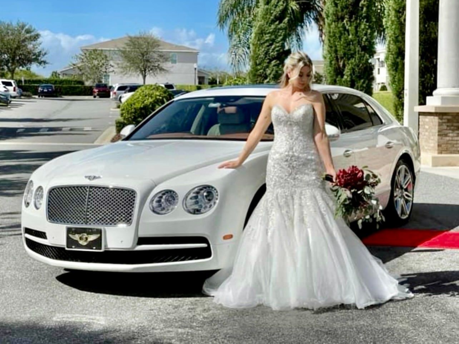A person in a white wedding gown holding a red bouquet, posing next to a white luxury sedan parked on a sunny day.
