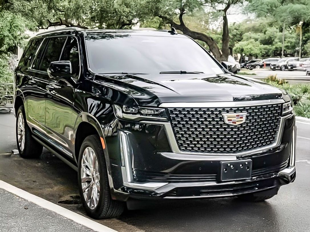 A black Cadillac Escalade SUV parked on a paved lot with trees and other cars in the background.