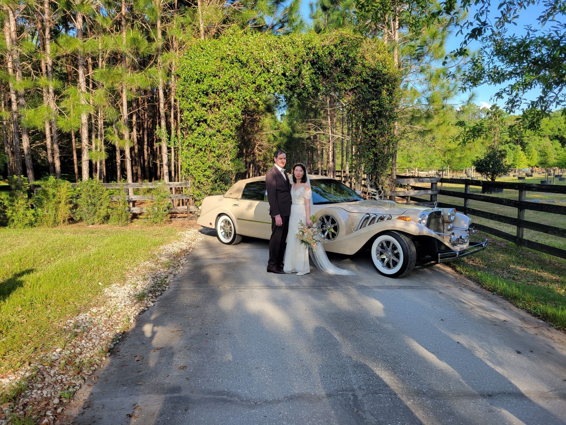 A couple stands beside a vintage cream-colored wedding car on a paved path lined with trees and a wooden fence.