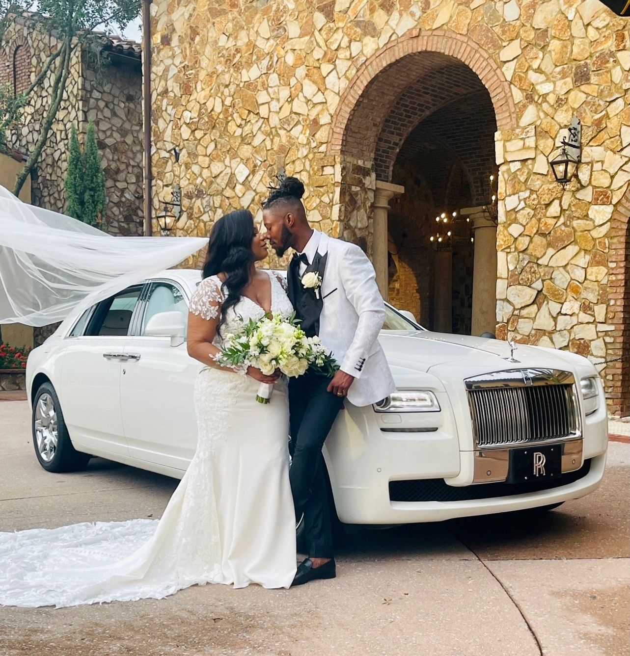 A bride and groom stand facing each other before a white luxury car in front of a stone building.
