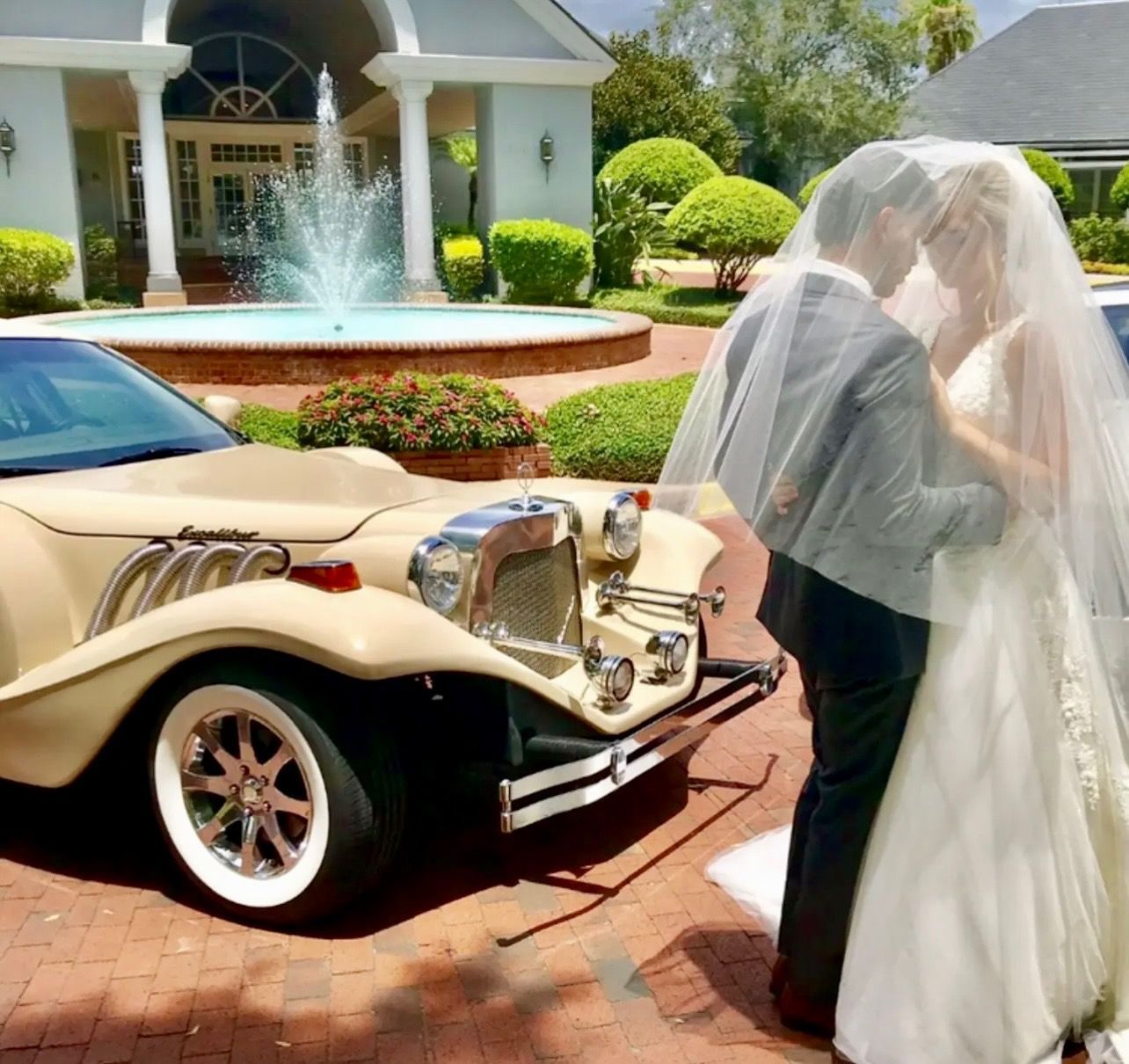 A bride and groom share an intimate moment under a veil next to a classic tan vintage car in front of a fountain.