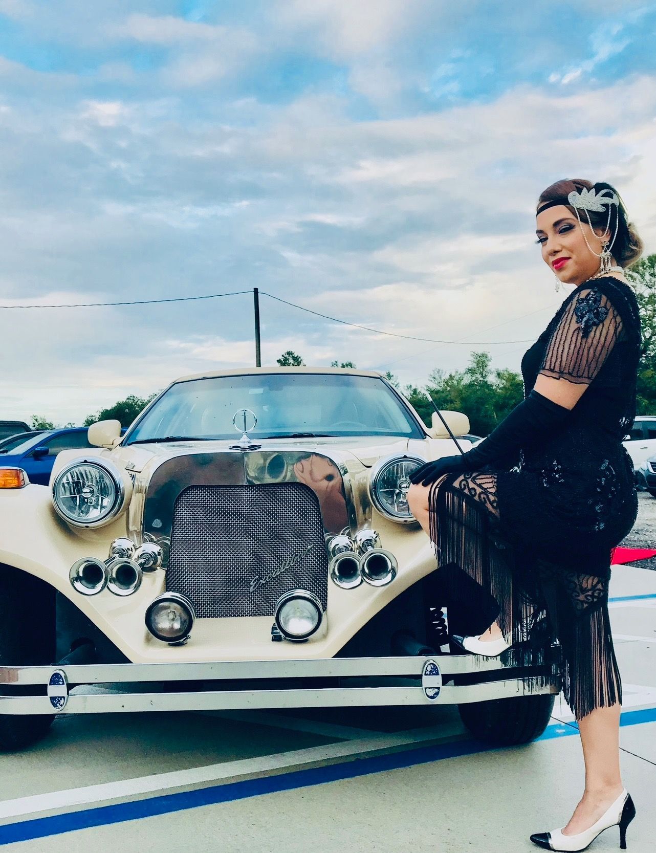A person dressed in a 1920s-style black flapper dress and headband poses next to a vintage-style cream convertible car.