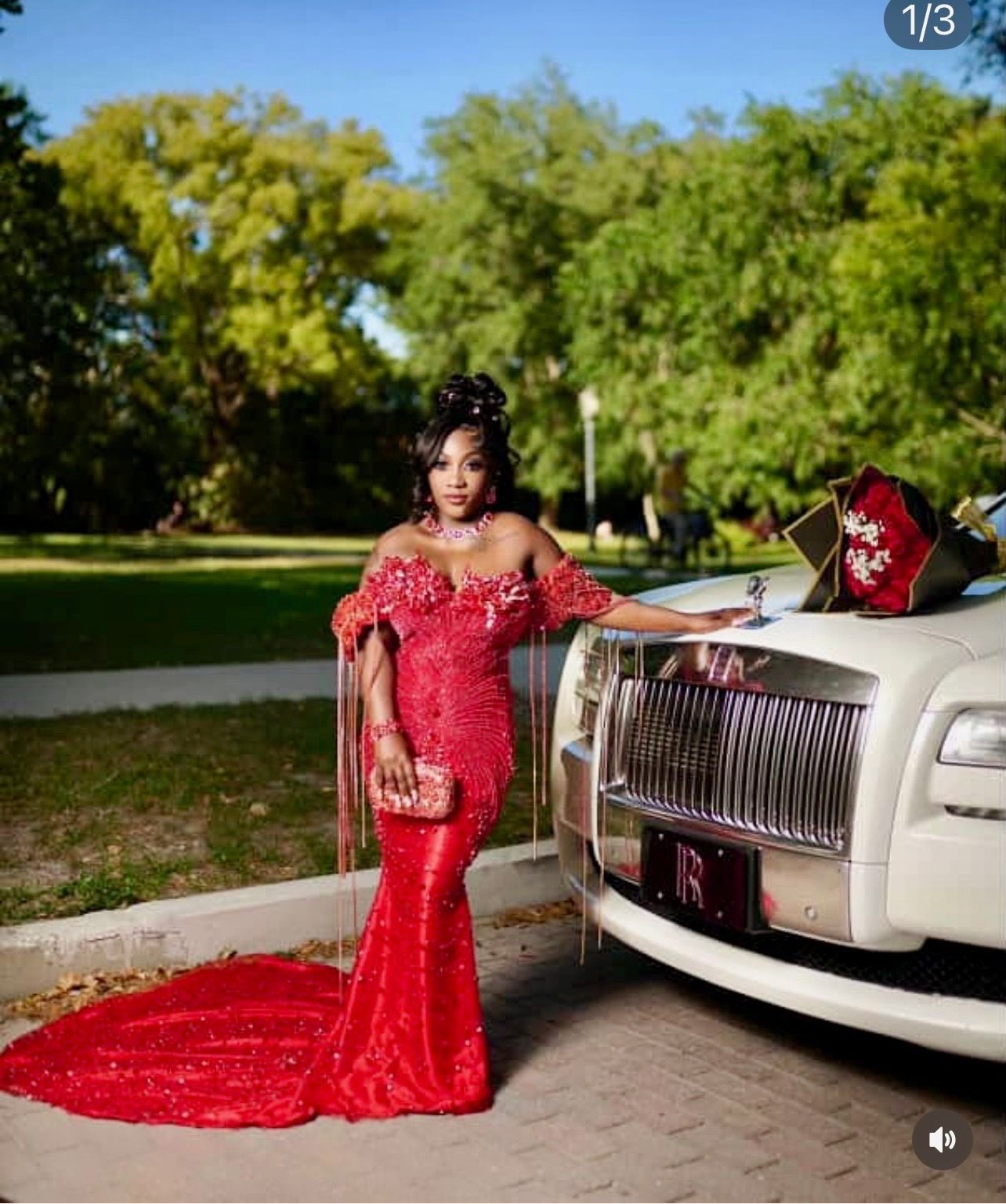 A person in a red, off-the-shoulder sequined gown leans against the front hood of a white luxury car in a park.