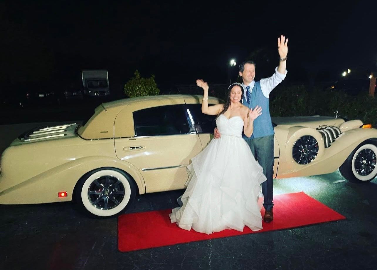 A bride and groom wave while standing on a red carpet next to a vintage cream-colored car at night.