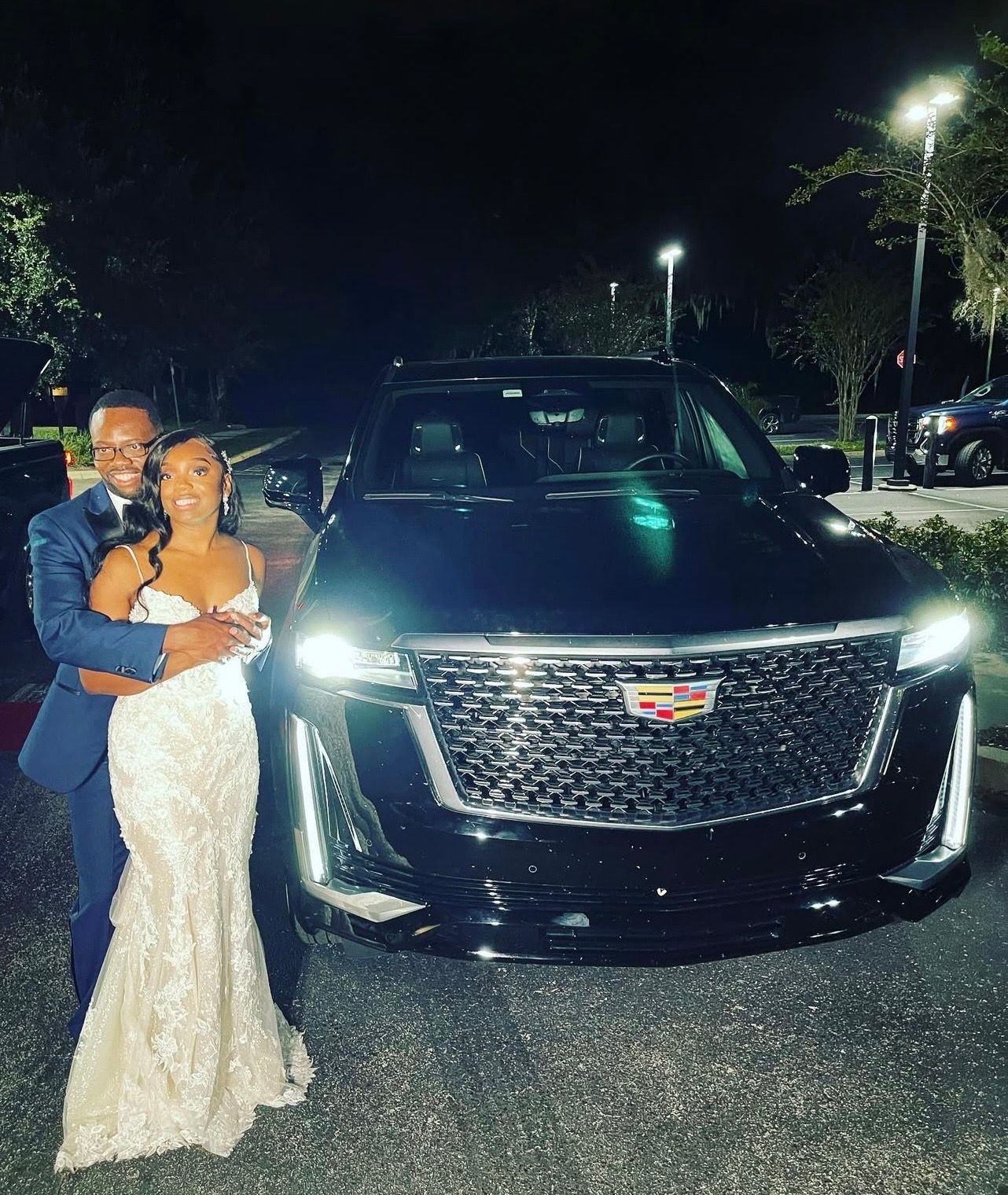 A couple in formal attire stands posing for a photo at night in front of a black Cadillac SUV with its headlights on.