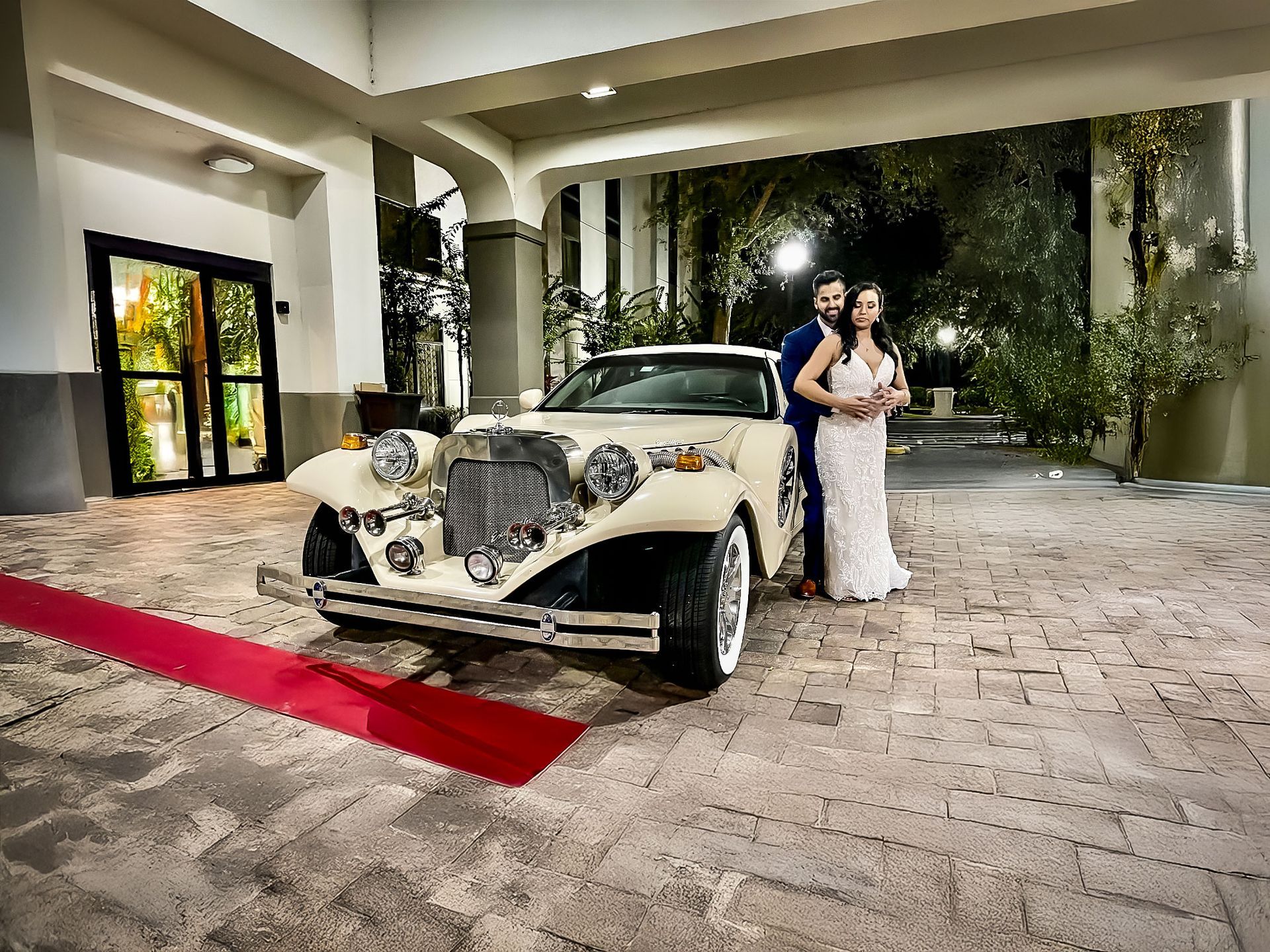 A couple stands next to a cream-colored vintage car on a paved driveway with a red carpet leading to an entrance.