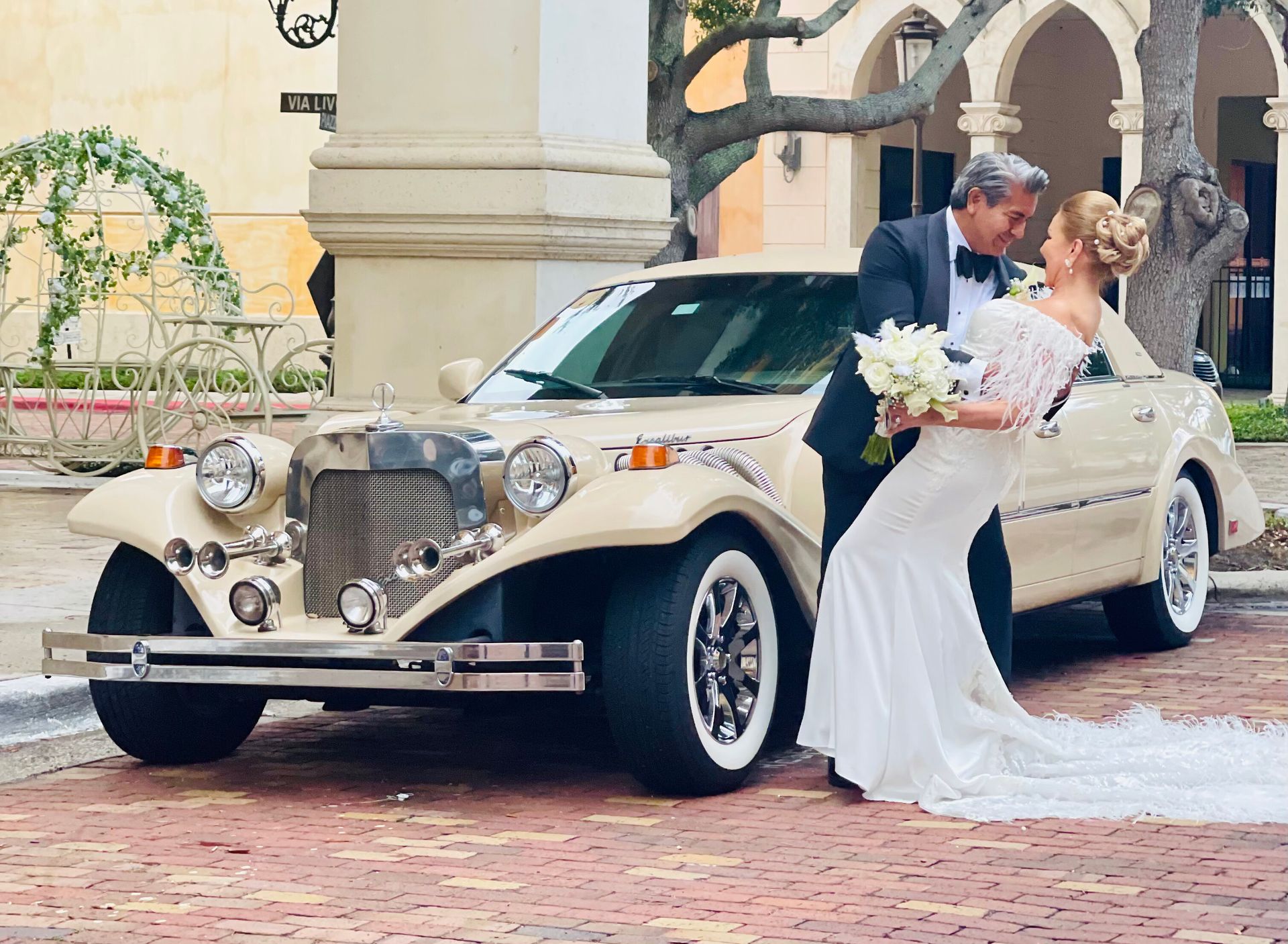 A couple in formal wedding attire embraces in front of a vintage, cream-colored convertible on a brick patio.